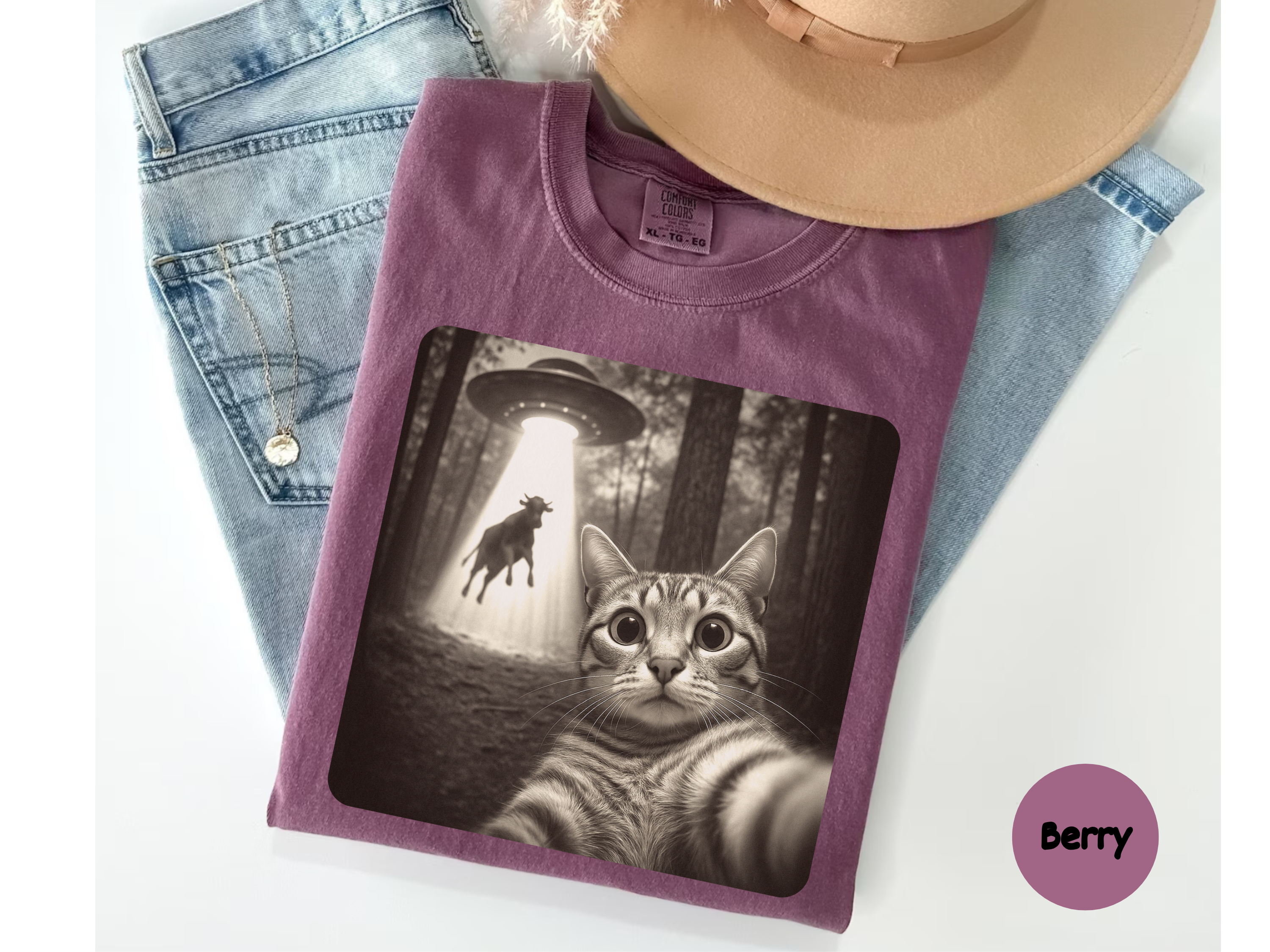 Purple t-shirt with a graphic of a cat in a forest and UFO design, placed on jeans.