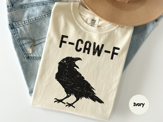 Beige t-shirt with a black crow silhouette and 'F-CAW-F' text, displayed with denim jeans and a beige hat.