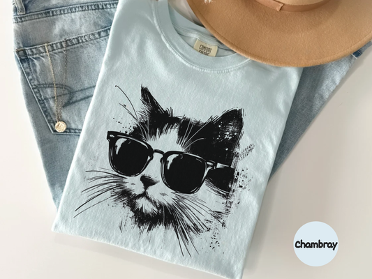 Light blue t-shirt with a black cat wearing sunglasses graphic, placed on a light surface with denim jeans and a beige hat.