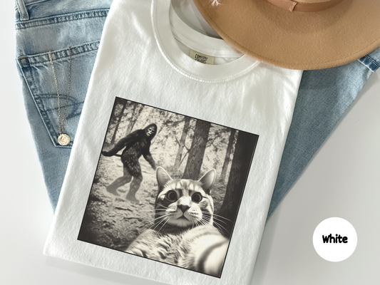 Close up of a humorous cat selfie graphic on a tee with Bigfoot walking behind in the woods combining viral meme style with cryptid folklore for conspiracy fans and animal lovers