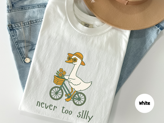Cute t shirt featuring a cartoon goose riding a bicycle with flowers and the phrase Never Too Silly on a black background perfect for animal lovers wholesome humor fans and cozy aesthetic outfits