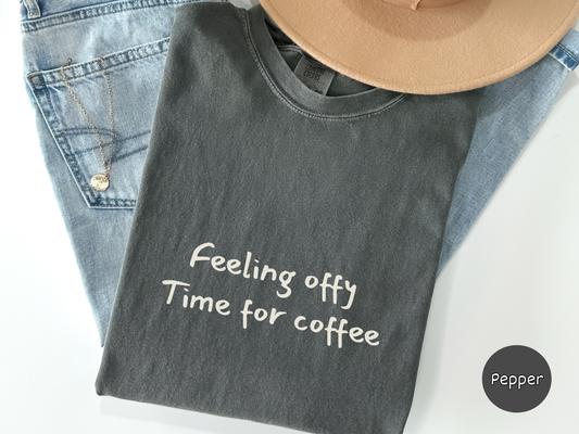 Close up of a black shirt with the words Feeling Offy Time For Coffee displayed in a simple typography style made for coffee addicts relatable humor fans and casual everyday wear