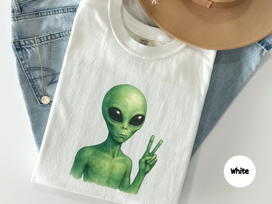Funny alien t shirt featuring green extraterrestrial holding up peace sign with large black eyes on dark background