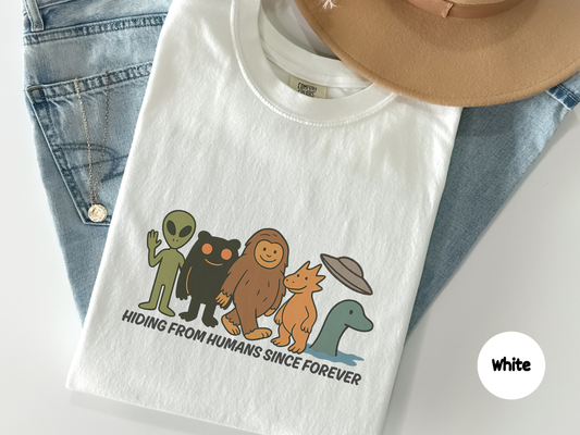 funny cryptid shirt illustration showing alien bigfoot mothman dogman loch ness monster and ufo standing together with text hiding from humans since forever