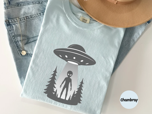 alien abduction ufo shirt graphic showing classic flying saucer hovering over forest while gray alien stands in bright beam of light on black background