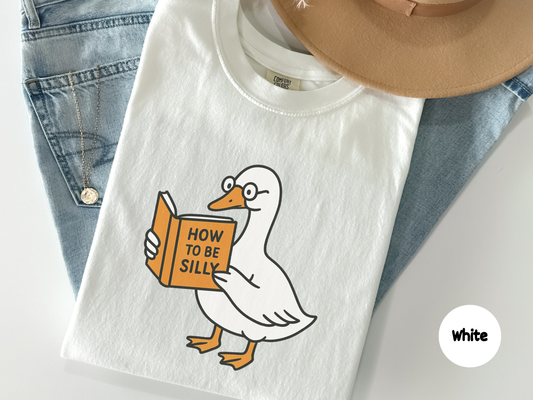Funny t shirt featuring a cartoon goose wearing glasses and reading a book titled How to Be Silly against a dark background perfect for book lovers animal fans and lighthearted humor